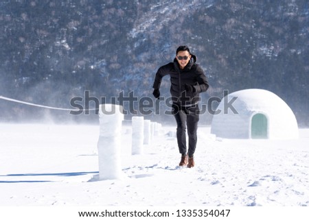 Young Asian man happiness smiling in winter clothes on frozen lake with ice Eskimo igloo.Funny male running on ice in cheerful joyful.Beautiful landscape in frost day.Travel Vacation Holiday Concept.