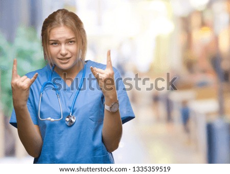 Young caucasian doctor woman wearing surgeon uniform over isolated background shouting with crazy expression doing rock symbol with hands up. Music star. Heavy concept.