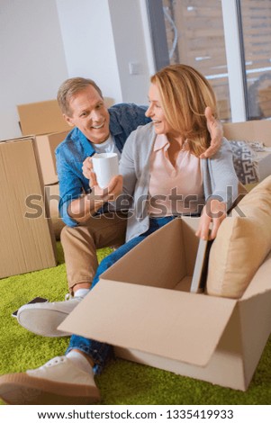 cheerful couple unpacking cardboard box while sitting on green carpet
