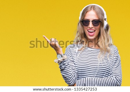 Beautiful young blonde woman wearing headphones and sunglasses over isolated background with a big smile on face, pointing with hand and finger to the side looking at the camera.