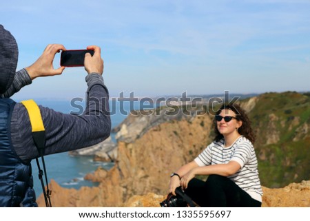 A man photographs a girl with a camera on the phone. Cabo da Roca, Portugal. Cropped shot, horizontal, front view, perspective view. The concept of family and leisure.