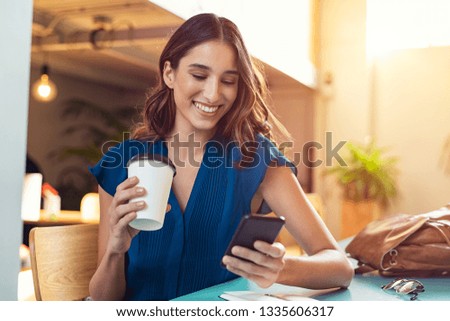 Young beautiful woman holding coffee paper cup and looking at smartphone while sitting at cafeteria. Happy university student girl using mobile phone. Businesswoman drinking coffee and smiling. Royalty-Free Stock Photo #1335606317
