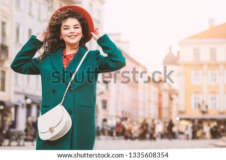 Young beautiful happy smiling lady wearing orange hat, green coat, with white cross body bag posing in street of European city. Copy, empty space for text Royalty-Free Stock Photo #1335608354