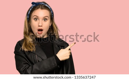 Teenager girl with leather jacket surprised and pointing side on isolated pink background