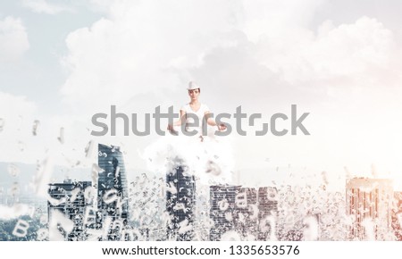 Woman in white clothing keeping eyes closed and looking concentrated while meditating on cloud among flying letters with cityscape view on background.