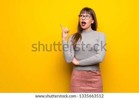 Woman with glasses over yellow wall thinking an idea pointing the finger up
