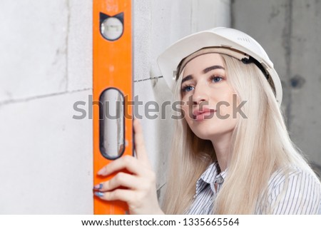 Closeup blonde girl designer foreman in white construction helmet measures the grey wall of sibit orange building level in house under construction. Сoncept female labor, repair shoot