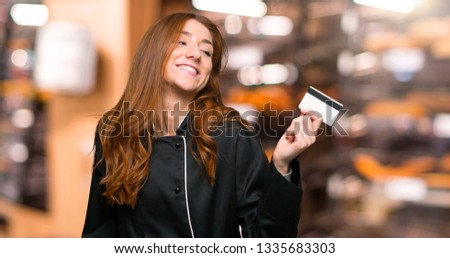Young redhead chef woman holding a credit card and thinking in the bakery