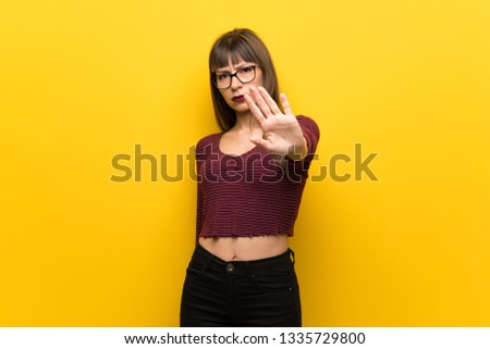 Woman with glasses over yellow wall making stop gesture denying a situation that thinks wrong