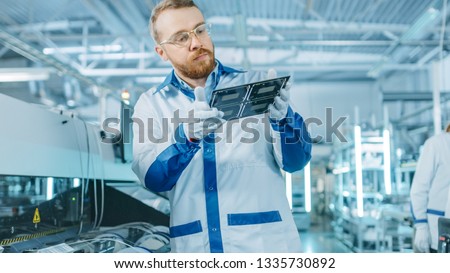 High-Tech Factory: Quality Control Inspector Checks Electronic Printed Circuit Board it for Damages. In the Background Assembly Line for PCB with Surface Mount Pick and Place Technology. Royalty-Free Stock Photo #1335730892