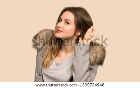 Teenager girl with coat listening to something by putting hand on the ear on isolated yellow background