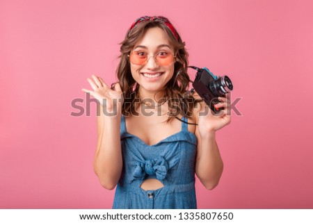attractive smiling woman with funny emotional face expression with vintage camera in denim dress and sunglasses isolated on pink background, traveler on vacation, summer fashion style, excited tourist