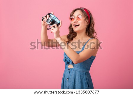 attractive smiling woman taking photo on vintage camera wearing denim dress and sunglasses, isolated on pink background, traveler on vacation, summer fashion style, excited tourist