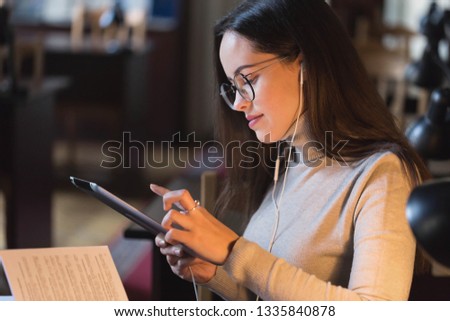 Clever student woman wearing glasses standing with the tablet and searching something on it while listening music