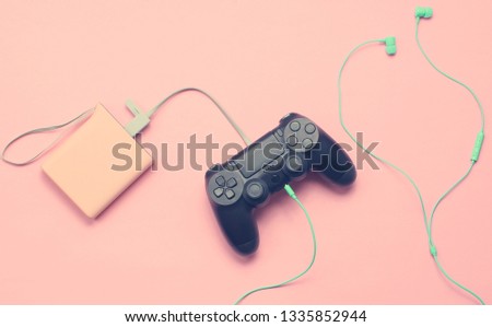Gamepad is charging from the power bank, earphones on pink background, Modern devices, top view

