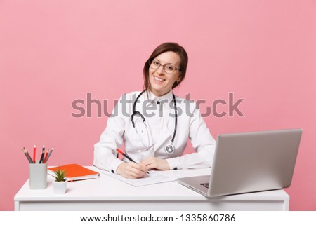 Beautiful female doctor sits at desk works on computer with medical document in hospital isolated on pastel pink background. Woman in medical gown glasses stethoscope. Healthcare medicine concept