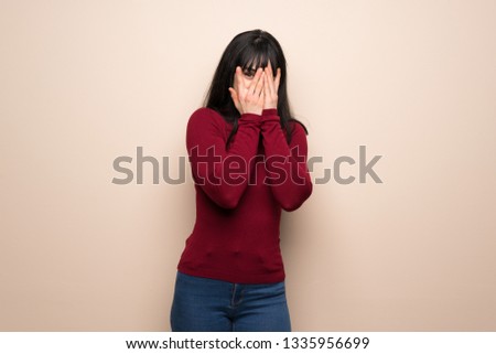 Young woman with red turtleneck covering eyes by hands and looking through the fingers