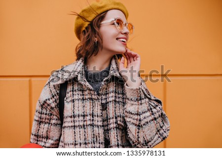 Portrait of model in stylish sunglasses, beret and checkered jacket. Young girl with smile looks away against background of orange wall