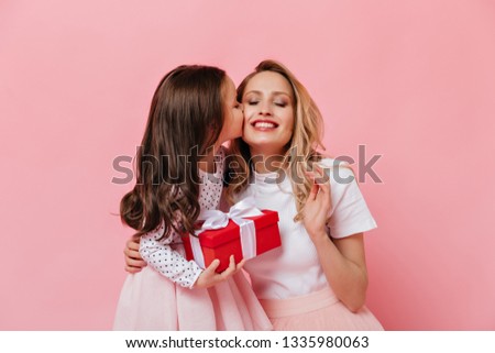Close-up portrait of curly female child kissing mom. Girl makes surprise to her older sister