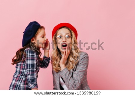Female child tells secret to adult surprised woman. Photo of mother and daughter in checkered jackets and multi-colored berets
