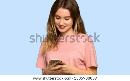 Teenager girl with pink sweater sending a message with the mobile on isolated blue background