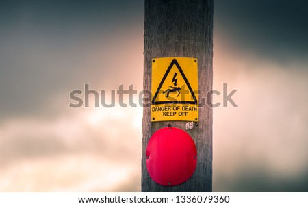 Danger of Death due to electrocution warning sign attached to a weathered wooden electricity pylon. Dark storm clouds can be seen in the background. 