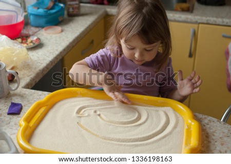 Little girl draws a finger on a tray with sand in the kitchen, make fine motor skills with drawing