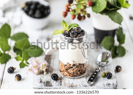 Handmade granola with white natural yogurt with blackberries in a glass transparent jar, flowers and leaves on a white wooden background.