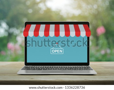 Modern laptop computer with online shopping store graphic and open sign on wooden table over blur pink flower and tree in garden, Business internet shop online concept