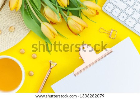 Office desk workplace with teae cup, clipboard, tulip bouquet, hat and keyboard on yellow background. Top view with copy space