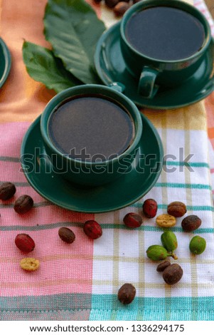 Cup with black coffee served outside with raw green, mature red and roasted coffee beans, decorated with green leaves from coffee plant close up