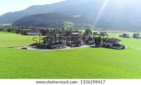 Aerial photo of Alps region showing some alpine apartments located at fresh green grass meadow during beautiful summers day with bright sunlight and mountain range in background