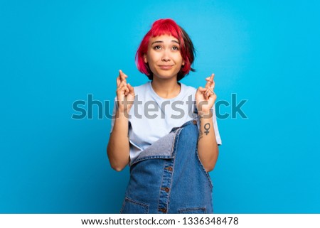 Young woman with pink hair over blue wall with fingers crossing and wishing the best