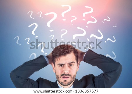 Portrait of confused young businessman with beard wearing dark suit and looking forward standing near blue wall with question marks drawn on it