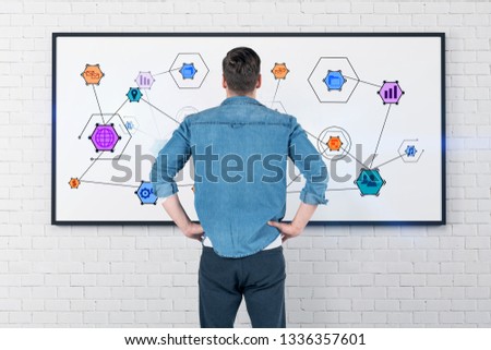 Rear view of caucasian young man in jeans shirt and gray pants looking at whiteboard with colorful internet icons on it.