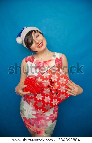 plump woman with short black hair and santa hat standing in white dress with gift boxes and so excited on blue studio background