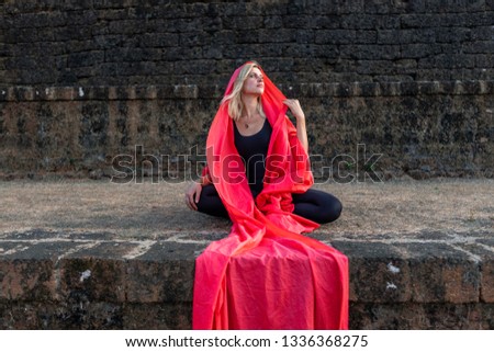 Blonde young yoga woman practicing yoga concept, sitting after class on the background of black old Fort in India, working out, head covered with red scarf, silhouette, black background. Healthy lifes