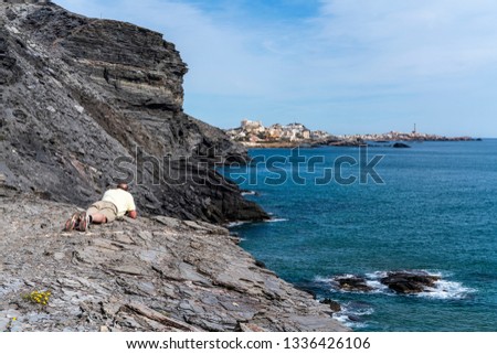 Man in yellow shirt and shorts lies face down over the edge of a grey sandstone cliff to photograph the rocky shore of Calblanque, Murcia, Spain. The town of Cabo de Palos in the distance.