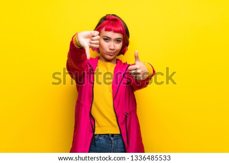 Young woman with pink hair over yellow wall making good-bad sign. Undecided between yes or not