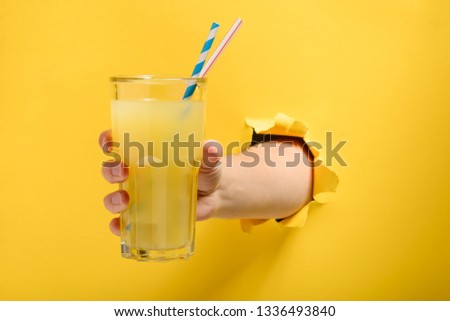 Hand giving a glass of cocktail with straws through torn yellow paper wall. Summer drinks to get cool and relax.