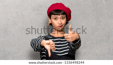 Young woman with beret making good-bad sign. Undecided between yes or not over textured wall