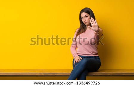 Young woman sitting on table making stop gesture denying a situation that thinks wrong