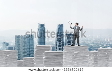 Businessman in suit standing on pile of documents with speaker in hand with cityscape on background. Mixed media.
