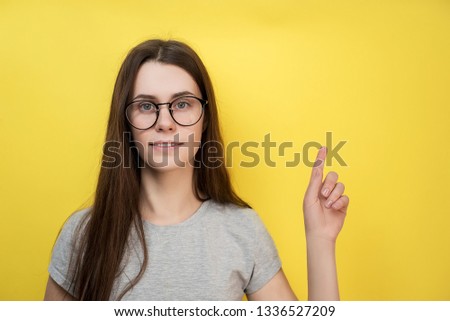Pleased lovely young female in glasses, indicates with fore finger upstairs, shows blank space, wears gray T-shirt. Successful woman distance worker gestures indoor