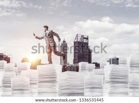 Businessman in suit running with phone in hand on pile of documents with sunlight and cityscape on background. Mixed media.
