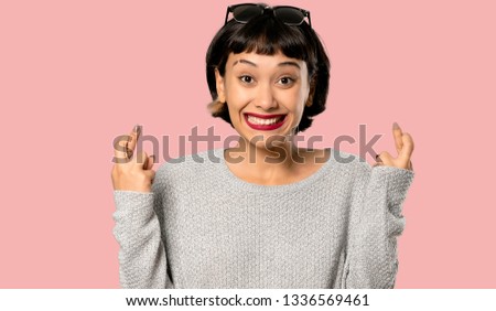 Young woman with short hair with fingers crossing and wishing the best on isolated pink background