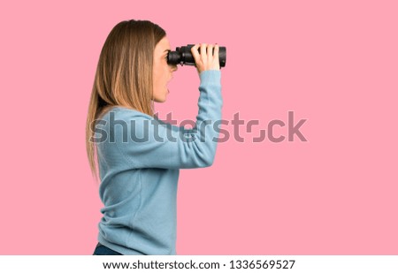 Blonde woman with blue shirt and looking in the distance with binoculars on isolated pink background