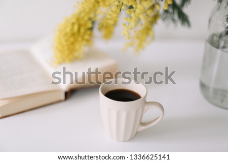 Cup of coffee and a book on white desk with yellow flowers bouquet mimosa. Planning and design concept. Workplace. Instagram feminine flat lay. Top view. Cozy breakfast.  Easter, spring concept