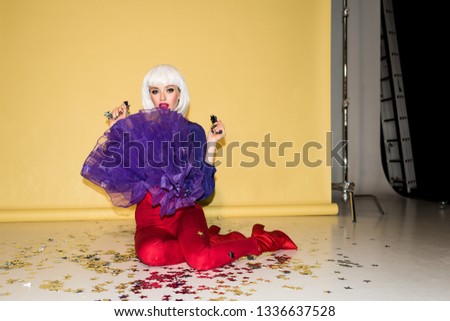 Gorgeous girl in wig sitting on floor and holding confetti on yellow background