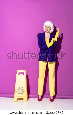 Girl in wig standing near wet floor sign and putting on rubber gloves on purple background
